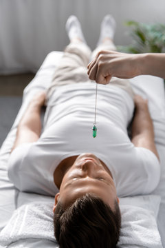 cropped view of hypnotist standing near man on massage table and holding green stone