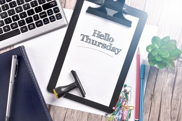 Keyboard with notebook and clipboard on wooden table with text HELLO THURSDAY