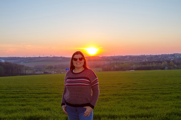 Lady poses outside at sunset for a portrait with the sun behind her. She is wearing sunglasses.
