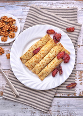 Pancakes, Walnut, Plums. Pancakes with filling on a white plate. Wood background. Rustic style. Rolled up pancakes. Wrapped thin pancakes. Homemade crepes, dried fruits, nuts.