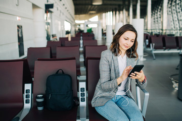 Young girl at the airport waiting for her departure. Scrolling smartphone in headphones. A cup of...