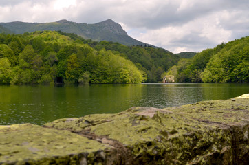 Green lake surrounded by a forest of green and yellow trees and a house at the foot of the lake. In the foreground a stone wall - Montseny National Park, Catalonia, Spain.
