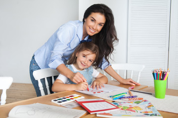 Smiling mother and daughter are preparing for school and engaged in drawing with pencils and paints