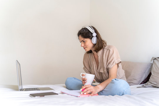 Young woman sitting on bed with headphones and laptop taking notes