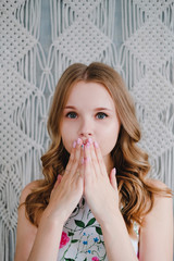 Portrait of a young cute woman covering mouth with her hands on background of macrame wall.