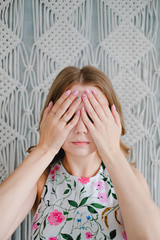 Portrait of a young woman covering her eyes with hands on background of macrame wall.
