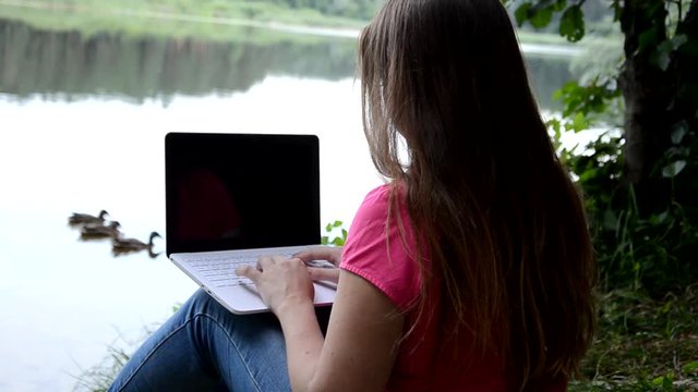 Cute Blonde girl sitting on the lake and working with laptop in summer or sping autumn day against beautiful landscape of northern lake and forest. Freelance, work and travel concept. call the phone