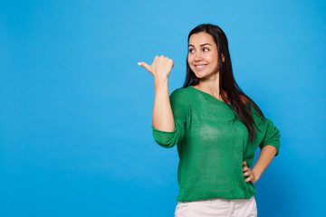 Fototapeta premium Portrait of smiling attractive young woman in green casual clothes looking and pointing thumb aside isolated on bright blue wall background in studio. People lifestyle concept. Mock up copy space.
