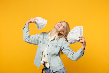 Portrait of joyful young woman in denim casual clothes hold fan of cash money in dollar banknotes isolated on yellow orange wall background in studio. People lifestyle concept. Mock up copy space.