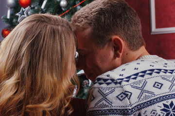 Couple in love sitting next to a Christmas tree, wearing warm sweaters, hugging and looking away from the camera towards the tree.