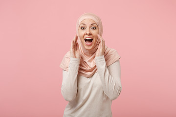 Excited young arabian muslim girl in hijab light clothes posing isolated on pink background in studio. People religious Islam lifestyle concept. Mock up copy space. Screaming with hands near mouth.