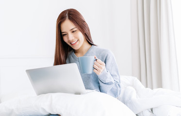 Happy young Asian woman in gray sweater relaxing watching videos or enjoying entertainment and holdig a coffee cup sitting on the bed at home in the winter