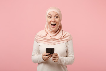 Surprised young arabian muslim woman in hijab light clothes posing isolated on pink background. People religious Islam lifestyle concept. Mock up copy space. Using mobile phone, typing sms message.