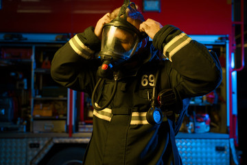 Photo of fireman putting on gas mask on background of fire engine at fire station.