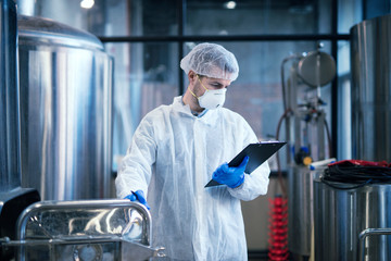 Technologist working in food processing factory checking quality and production. Industrial worker in white protective clothes holding checklist and reading results.
