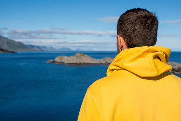 Fototapeta premium A young man in a yellow hoodie outdoor enjoy the northern beauty. Ocean and rocks landscape. Scenic view. Travel, adventure. Sense of freedom, lifestyle. Lofoten Islands, Norway. Summer in Scandinavia