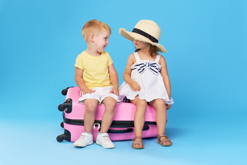 Happy kids sitting on colorful pink suitcase prepared for summer vacation. Young travelers. Little girl and boy, sister and brother, having fun isolated on blue background