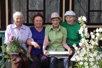 Four Russian elderly women, close friends, sit on a bench at the village house and rest. One senior is holding a