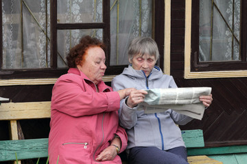 Two Russian women sit on a bench at the village house and actively discuss the news from the newspaper. Country life.