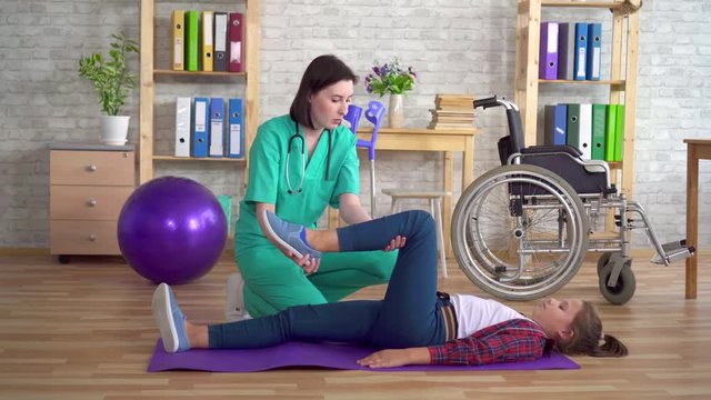 Physiotherapist in the rehabilitation center helps in warming up the legs of a teenage girl in rehabilitation after a spinal injury