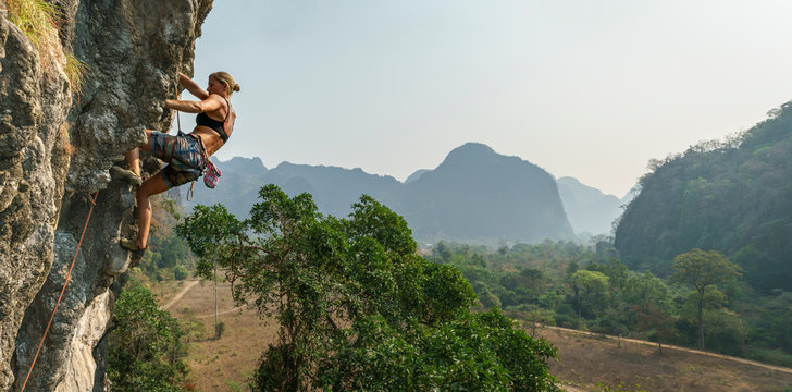 Female rock climber high on wall with trees and mountains behind, Asia