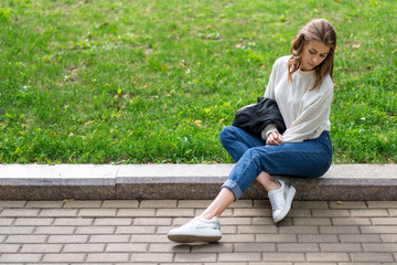 beautiful slender girl is resting in city park. Caucasian girl posing sitting on curb sidewalk. Lifestyle, pullover, jeans, sneakers, jacket. Cool posing while sitting on grass in trendy look.