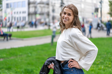 young beautiful european girl long flowing hair walks in city park in great mood. Smile, white teeth. Wearing pullover. In hands black leather jacket. Happy girl 25 years old. Rainy weather.