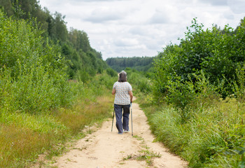 Elderly senior woman practicing nordic walking in the forest, healthy lifestyles in old age. An old pensioner walks with sticks in the woods. Sport walking in a summer sunny day.