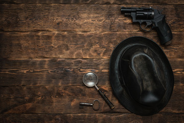 Detective or spy agent wooden table flat lay background with copy space. Handgun, black leather hat, magnifying glass and a rusty key on the work table.