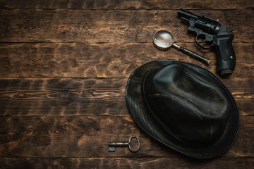 Detective or spy agent wooden table flat lay background with copy space. Handgun, black leather hat, magnifying glass and a rusty key on the work table.