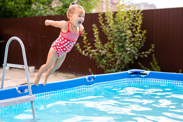 Happy little girl in red swimsuit jumping into outdoor swimming pool at home. Baby girl learning to...