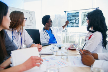 Young Afro American doctor shows mri patient's result and discusses it with the team. International group of five medical workers have a meeting at conference room in hospital.