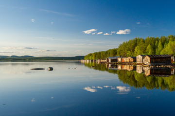 Fototapeta na wymiar Old wooden boat sheds on Lake Kovdozero in the Murmansk region, Zelenoborsky village. The reflection in the water of trees and boat garages