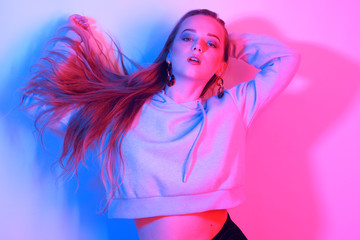 Fashion young elegant girl. Colored neon background, studio shot. Beautiful brunette woman. Hipster girl dancing in neon. Woman with stylish hair and red lips. Girl in a sweater and long hair.