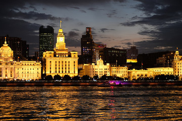 Obraz na płótnie Canvas Shining golden western buildings under sunset clouds sky. River waves and reflection. Beautiful evening scene of the bund in Shanghai Puxi, on the bank of Huang Pu River. 