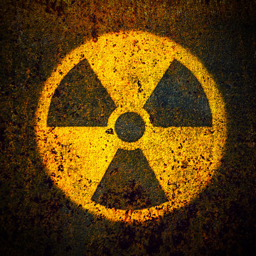 Round yellow radioactive (ionizing radiation) danger symbol painted on a massive rusty metal wall plate with dark rustic grungy texture background in square format.