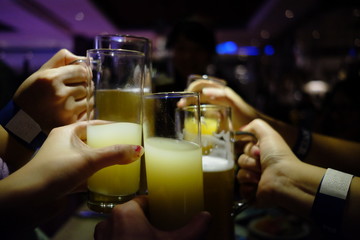 cheers! close up several hands of group of people raise glasses with yellow juice and wine champagne, cheering under bar light in party. dark blur background. cooperation teamwork concept