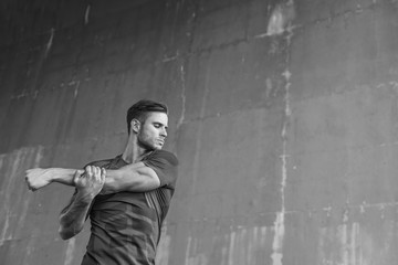 Black and white. Athlete doing stretching warmup exercise. Male fitness model in fashion sportswear in city. Runner sprinting on urban gray background.