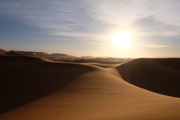 Fototapeta na wymiar wild brown Sahara desert sand dunes at sunset. Strong shining sun with white cloud. footsteps and shadows. Saharan, sandy near Merzouga in Morocco