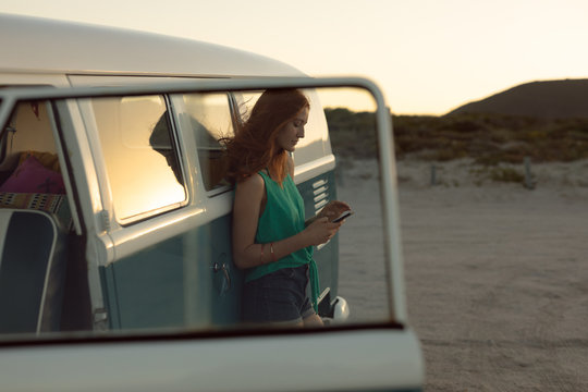 Beautiful woman leaning on camper van and using mobile phone at beach