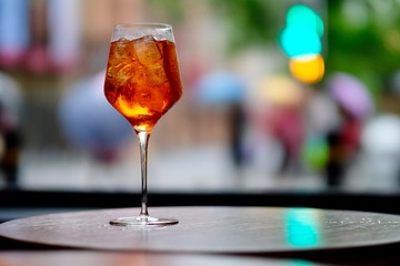 close up one wine glass of orange color cocktail on table, ice cubes and lemon slice inside. Defocused city lights and pedestrians with umbrellas as background