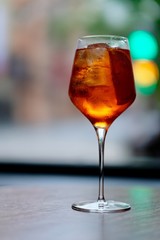 close up one wine glass of orange color cocktail on table , ice cubes and lemon slice inside. Defocused city lights background