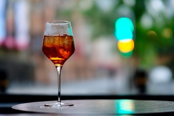 close up one wine glass of orange color cocktail on table , ice cubes and lemon slice inside. Defocused city lights background