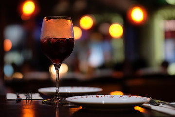 close up a glass of red cocktail on table. Colorful defocused illuminations, plates and fork. Dark background.