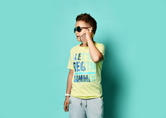 Portrait of a cool young teen guy talking on a mobile phone in summer style clothes in sunglasses, on a blue background.