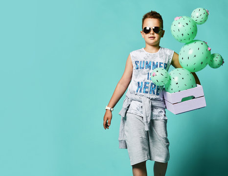 Cheerful boy boy teenager in summer lunch shorts and tank top holding green cacti-shaped balloons in a box.