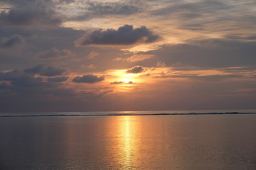 Naklejka premium Burning rising sun above wide sea horizon. Gray cloud skyline. Bright yellow sunlight reflection on ocean surface. Sunrise in Maldives