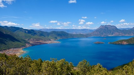 Fototapeta na wymiar deep blue lake surrounded by green mountains. Green trees foreground. Sunny blue sky with white clouds. high angle view of Lugu lake in Yunnan China