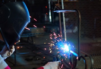 A Indian woman wearing a pink long sleeve jacket, welding mask and protective gloves, welding a metal chair, with sparks
