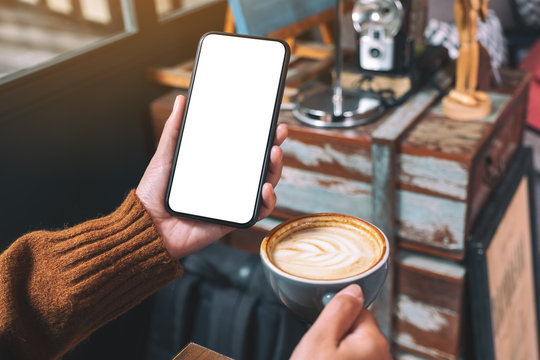 Mockup image of a woman holding black mobile phone with blank desktop screen while drinking coffee in cafe